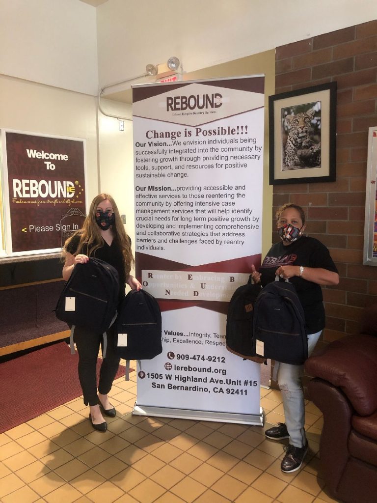 Two women hold several black backpacks each as they stand on either side of a sign for IE Rebound.