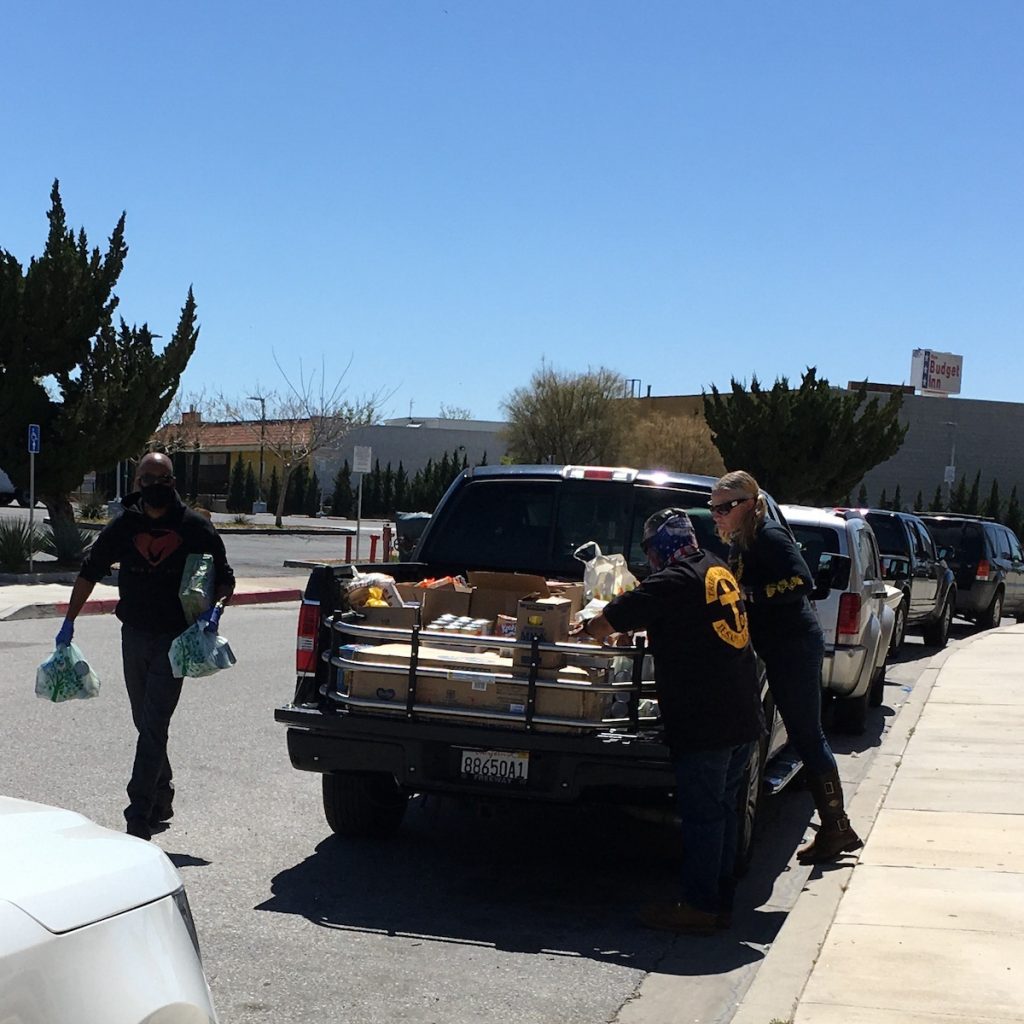 Two people stand by a pickup truck loaded with boxes of supplies. A third person approaches the truck with more supplies in bags.
