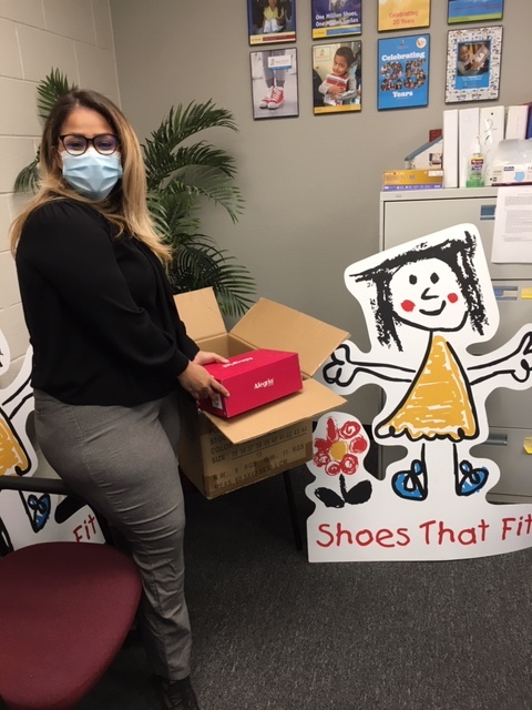 A woman in a black top and gray pants holds a shoe box next to a large sign with a picture of a girl that reads Shoes That Fit