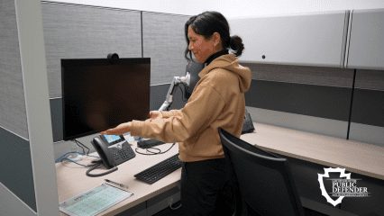 PSE adjusts monitor at cubicle desk with phone, calendar, and supplies. Public Defender logo in bottom right.