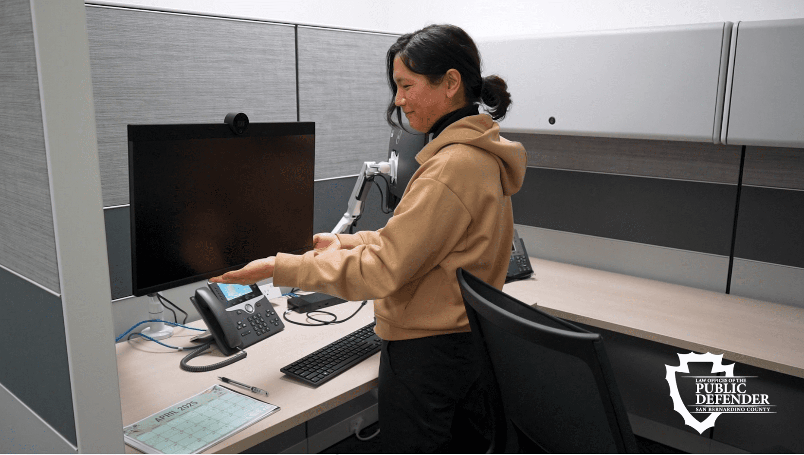 PSE adjusts monitor at cubicle desk with phone, calendar, and supplies. Public Defender logo in bottom right.