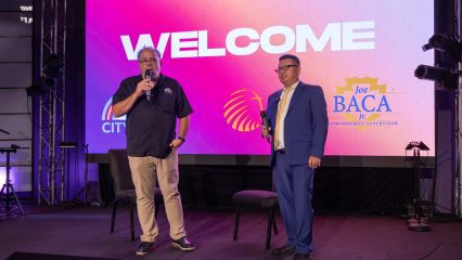 Two men with microphones speak on stage in front of a large screen displaying “WELCOME” and Joe Baca Jr.’s logo.
