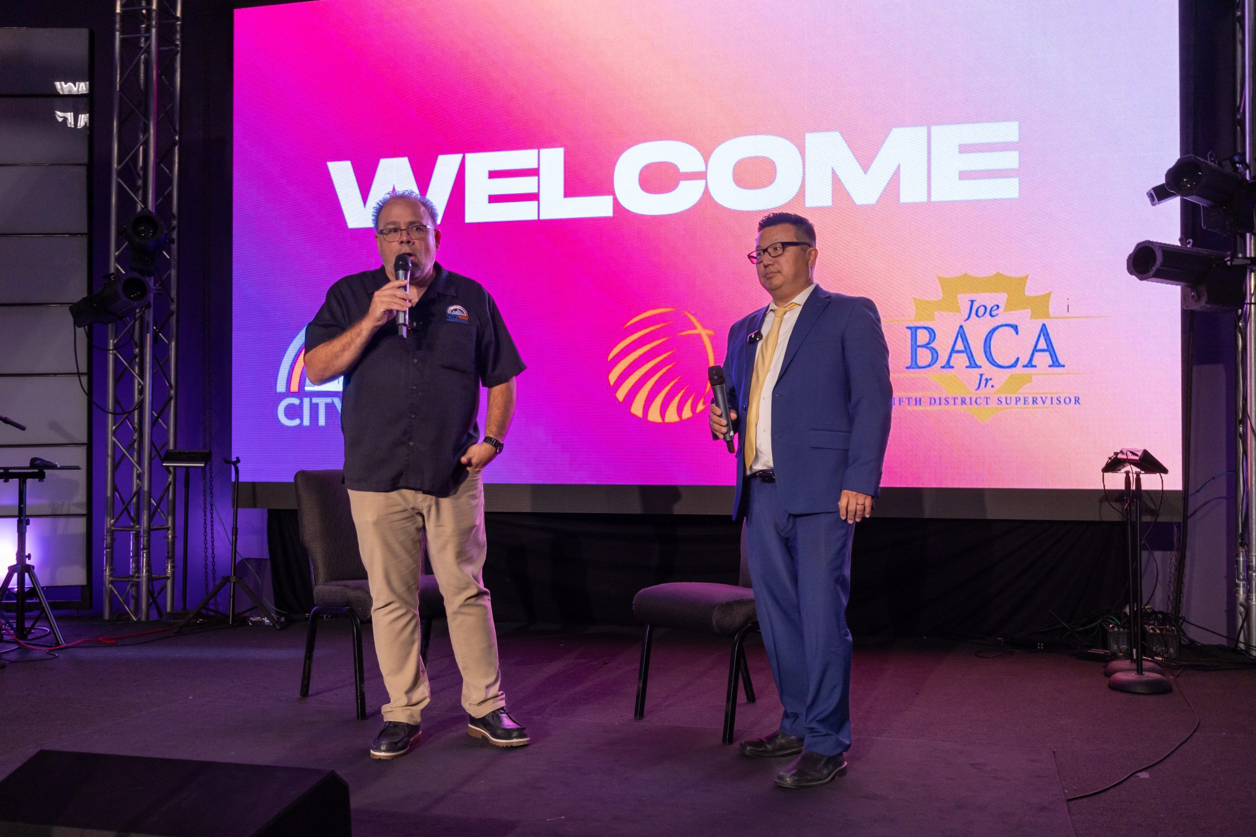 Two men with microphones speak on stage in front of a large screen displaying “WELCOME” and Joe Baca Jr.’s logo.
